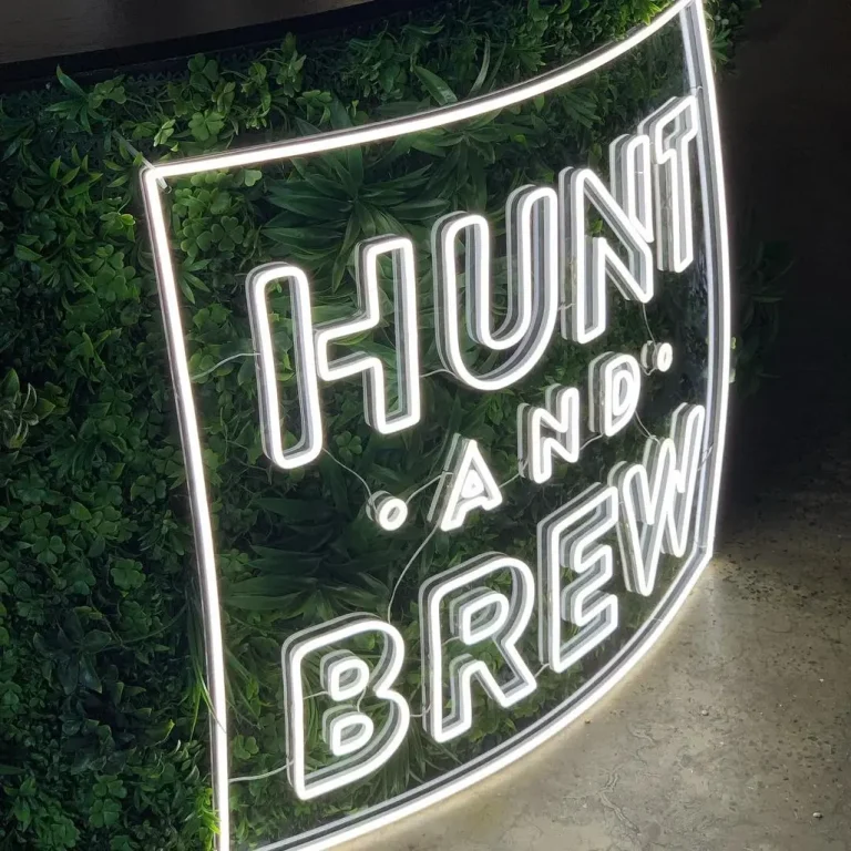 White LED neon sign displaying HUNT AND BREW text mounted on a green foliage wall perfect for a cafe or bar in Australia