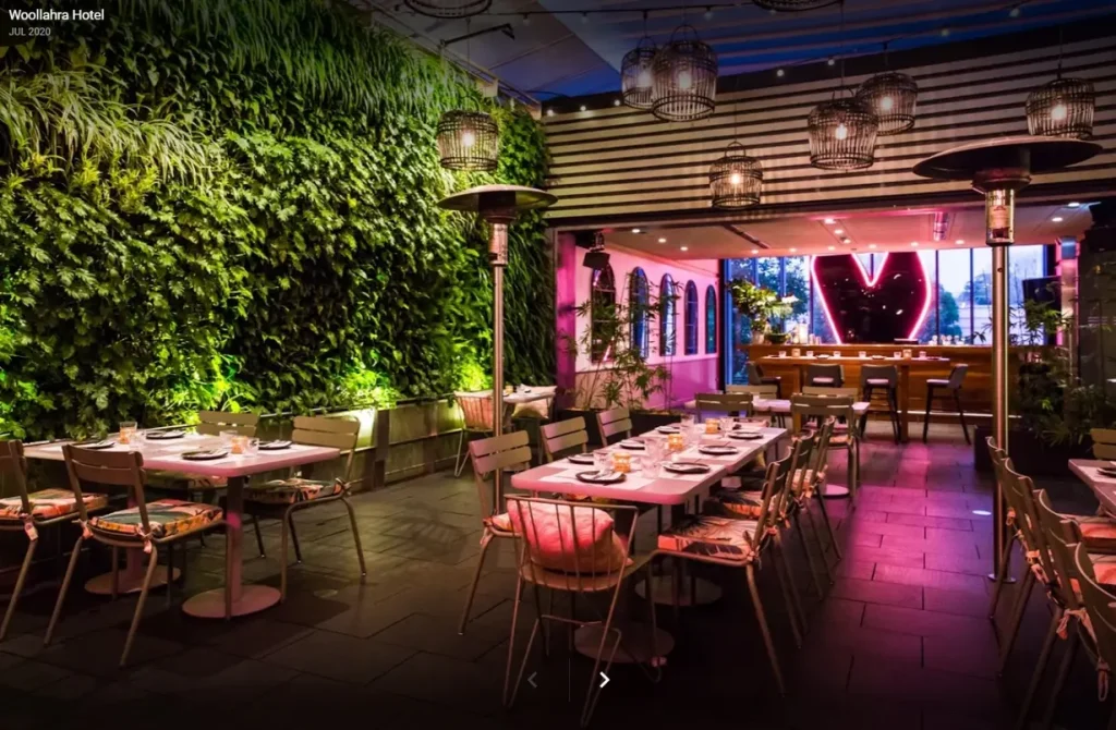 Woollahra Hotel Sydney Custom Pink LED Neon Heart Sign – Beyond Neon Signs Large pink LED neon heart sign glowing at the end of the Woollahra Hotel Sydney restaurant dining room with a green living wall, rattan pendant lights and set tables