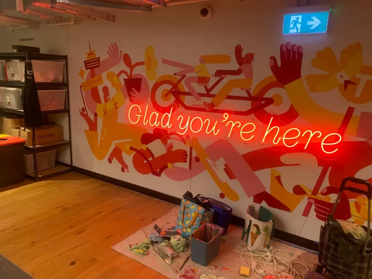Red LED neon sign reading Glad you're here in cursive script installed on a colourful illustrated mural wall inside an office co-working space with installation tools on the floor