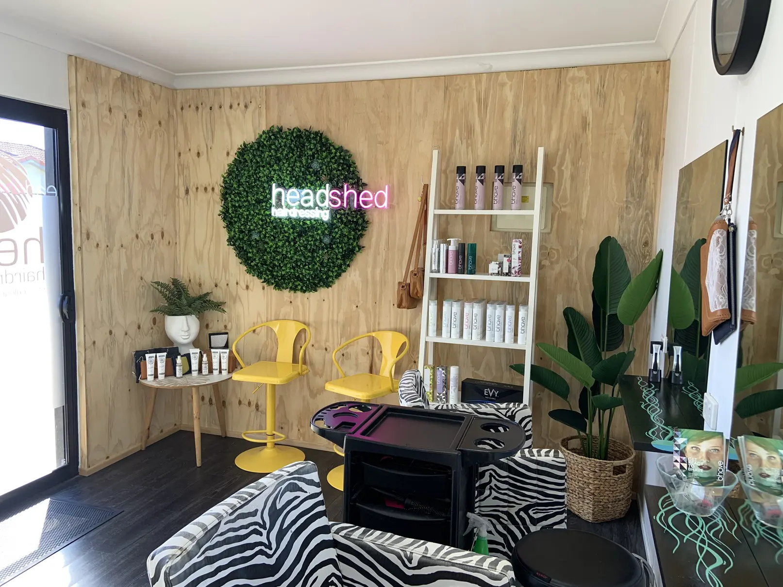 Custom LED neon sign made for Headshed Hairdressing installed on indoor salon wall above green hedge feature with wooden shelves and seating area showing easy plug and play installation