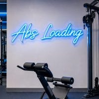 An 'Abs Loading' neon sign in Ice Blue, contour cut on clear acrylic, mounted on a dark moody training space wall.