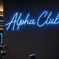 An Ice Blue 'Alpha Club' neon sign mounted on a dark, moody training space wall, illuminated with cinematic lighting.