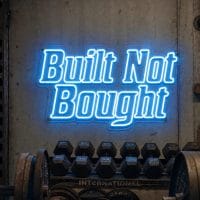 An Ice Blue 'Built Not Bought' neon sign mounted on a dark, textured concrete wall in an industrial-style gym, with barbells and weights visible in the foreground.