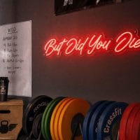 Red 'But Did You Die' neon sign mounted on a dark, textured wall in a moody training space with cinematic lighting.