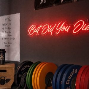 Red 'But Did You Die' neon sign mounted on a dark, textured wall in a moody training space with cinematic lighting.