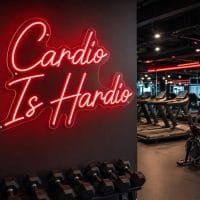 Red 'Cardio Is Hardio' neon sign, contour cut on clear acrylic backing with silver screws, mounted on a dark wall in an LED-lit fitness room.