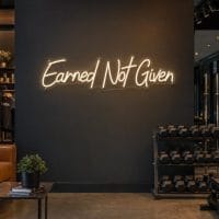 A warm white 'Earned Not Given' neon sign mounted on a dark moody training space wall, with cinematic lighting and a high-end commercial feel.