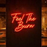 A red 'Feel The Burn' neon sign mounted on a dark wall in a moody training space, with cinematic lighting highlighting the sign's glow.