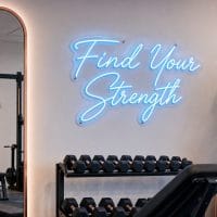 Ice Blue 'Find Your Strength' neon sign mounted on a dark concrete wall in an industrial gym setting, with gym equipment in the background.