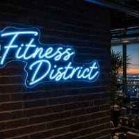 An Ice Blue 'Fitness District' neon sign mounted on a dark grey wall in a modern, LED-lit fitness room with exercise equipment in the background.