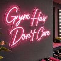 Pink "Gym Hair Don't Care" neon sign glowing brightly on a wall in a chic boutique Pilates studio, with natural light filtering in.