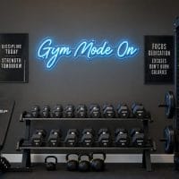 Ice Blue 'Gym Mode On' neon sign mounted on a dark, textured wall in a moody training space, with subtle backlighting.
