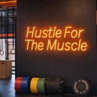 An orange 'Hustle For The Muscle' neon sign mounted on a dark, moody gym wall, with cinematic lighting highlighting the realistic LED glow.