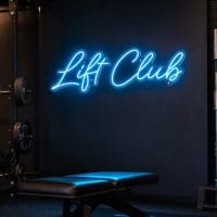 A bright Ice Blue 'Lift Club' neon sign mounted on a dark grey wall in a modern home gym with sleek black equipment and natural light.