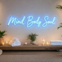An Ice Blue LED neon sign spelling "Mind Body Soul" mounted on a clear acrylic backing with silver screws, set against a serene wellness lounge background with soft, ambient lighting.