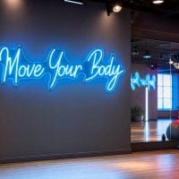 An Ice Blue 'Move Your Body' LED neon sign mounted on a dark grey wall in a modern home gym setting, with exercise equipment visible in the background.