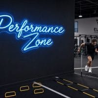 An Ice Blue 'Performance Zone' neon sign mounted on a dark, textured wall in a modern home gym with weights and exercise equipment visible.