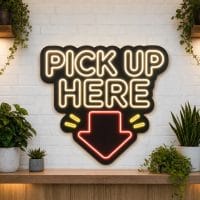 Pick Up Here LED neon sign on black acrylic backing mounted on white brick wall