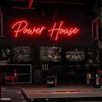 A red 'Power House' neon sign mounted on a dark concrete wall in an industrial-style gym, with gym equipment visible in the background.