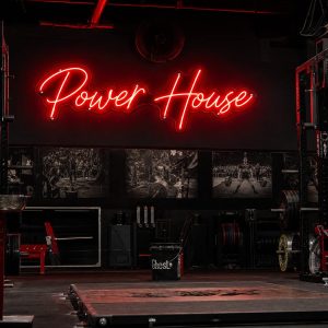 A red 'Power House' neon sign mounted on a dark concrete wall in an industrial-style gym, with gym equipment visible in the background.