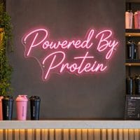 Pink 'Powered By Protein' neon sign mounted on a dark wall in a modern home gym setting, with gym equipment visible in the background.