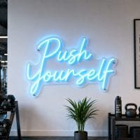 Ice Blue 'Push Yourself' neon sign mounted on a dark grey wall in a modern home gym with exercise equipment visible in the background, cinematic lighting.