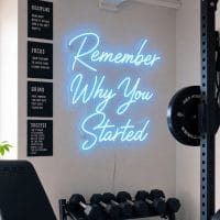 Ice Blue 'Remember Why You Started' neon sign mounted on a dark grey wall in a modern home gym with natural light, weights, and a yoga mat visible.