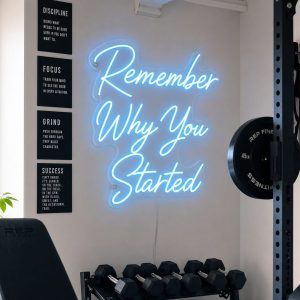 Ice Blue 'Remember Why You Started' neon sign mounted on a dark grey wall in a modern home gym with natural light, weights, and a yoga mat visible.