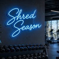 Ice Blue 'Shred Season' neon sign mounted on a dark wall in an LED-lit fitness room, with cinematic lighting and a realistic glow.
