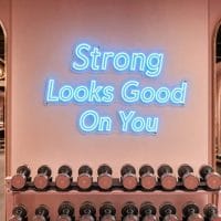 Ice Blue 'Strong Looks Good On You' neon sign mounted on a dark moody training space wall, with gym equipment subtly visible in the background.