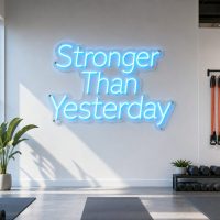 Ice Blue 'Stronger Than Yesterday' neon sign mounted on a dark grey concrete wall in an industrial gym setting, with gym equipment in the background.
