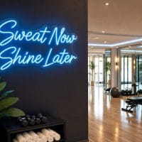 Ice Blue 'Sweat Now Shine Later' neon sign mounted on a dark wall in an LED-lit fitness room, with gym equipment in the background.