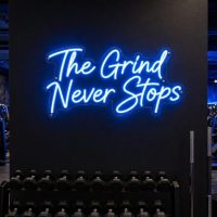 Ice Blue 'The Grind Never Stops' neon sign mounted on a dark, textured wall in a moody training space, with cinematic lighting highlighting the sign.