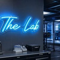 Ice Blue 'The Lab' neon sign mounted on a concrete industrial gym wall, with weights and equipment in the background.
