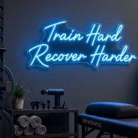 An ice blue 'Train Hard Recover Harder' neon sign mounted on a light grey wall in a modern recovery spa room, with soft, ambient lighting.