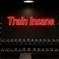 Red 'Train Insane' neon sign mounted on a dark moody training space wall, with gym equipment in the background.