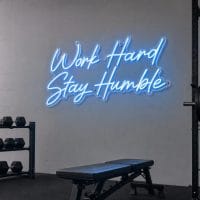 Ice Blue 'Work Hard Stay Humble' neon sign mounted on a dark concrete wall in an industrial gym setting, with silver screws visible.